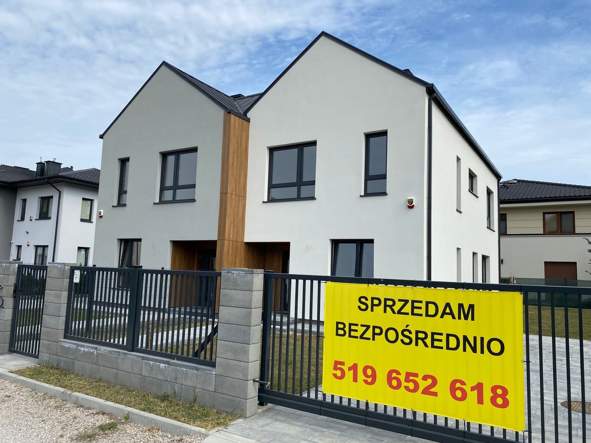 Bliźniak od ulicy za ogrodzeniem: biały tynk, drewniane wnęki wejściowe, żółty baner sprzedażowy na bramie